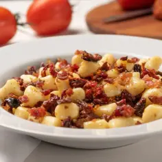 Pyszne gnocchi z boczkiem i suszonymi pomidorami na białym talerzu, w tle pomidory.