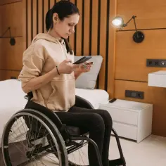 Kobieta na wózku inwalidzkim, zmagająca się z miopatią, korzysta z telefonu i karty płatniczej w hotelowym pokoju.