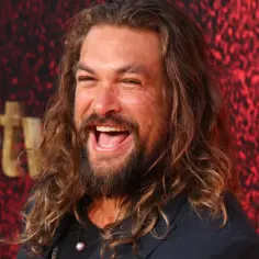 Jason Momoa śmieje się radośnie na tle czerwonej ściany z logo Apple TV+. Gwiazdor znany z wielu filmów, seriali i programów.