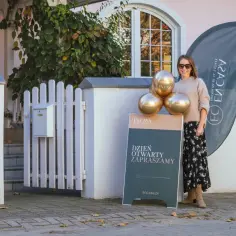Uśmiechnięta kobieta zaprasza na dni otwarte. Złote balony i tablica z napisem "Dzień Otwarty" informują o wydarzeniu.