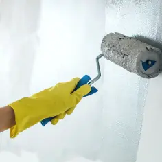 Ręka w żółtej rękawicy maluje ścianę wałkiem do farby lateksowej, nadając jej nowy, jasny odcień.