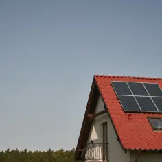 Panel fotowoltaiczny na czerwonym dachu domu, na tle błękitnego nieba.