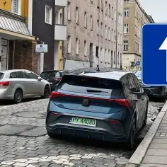 Znak D3 nakazuje jazdę prosto. Na ulicy zaparkowane są samochody, w tym ciemnoniebieski Cupra.