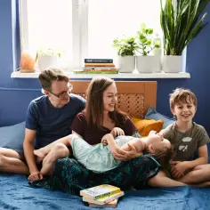 Rodzicielstwo bliskości w praktyce: rodzina śmieje się i przytula na łóżku. Mama tuli córeczkę, tata i starszy syn patrzą z uśmiechem.
