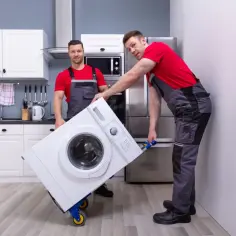 Dwóch mężczyzn w szarych ogrodniczkach i czerwonych koszulkach przewozi pralkę na wózku. Czy można przewozić pralkę na leżąco? Zapytaj fachowców.