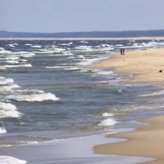 Wietrzny dzień nad ujściem Wisły. Dwie osoby spacerują po piaszczystej plaży, a fale rozbijają się o brzeg.