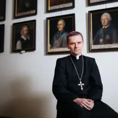Mężczyzna w stroju duchownym siedzi na tle ścian z portretami.