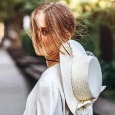 Kobieta z pięknymi bronde włosami, częściowo zasłoniętymi przez wiatr, odwraca głowę, trzymając elegancki kapelusz.