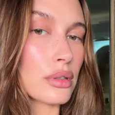 Zbliżenie na twarz Hailey Bieber z delikatnym makijażem i piegami. Jej skóra lśni, a usta są pełne.