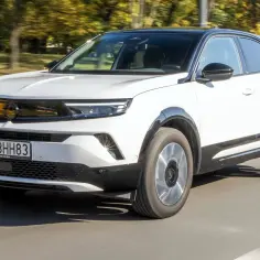 Biały Opel Mokka 2023 z czarnym dachem pędzi po drodze, otoczony jesiennymi drzewami.