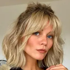 Kobieta z niebieskimi oczami i piegami, ma blond włosy z delikatnymi, **wispy bangs** i upięte w mały kok na czubku głowy.