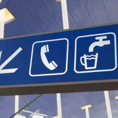 Niebieska tablica z białymi piktogramami: strzałka w lewo, słuchawka telefonu, kran z wodą do kubka. Czy na lotnisku w Katowicach można mieć wodę? Tak, jest punkt z wodą.