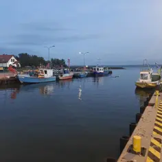 Spokojny wieczór w Krynicy Morskiej port. Kilka łodzi rybackich zacumowanych przy nabrzeżu, woda odbija światła.