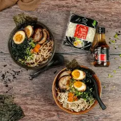 Dwa miski aromatycznego ramenu z jajkiem, mięsem i wodorostami. Obok makaron i sos, idealne składniki, jak zrobić ramen.