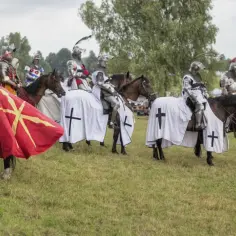 Rycerze w zbrojach i białych płaszczach z krzyżami, jak krzyżacy, przygotowują się do parady. Czerwony proporzec z żółtym symbolem.