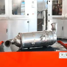 Metalowy element układu wydechowego, prawdopodobnie gpf, leży na pomarańczowej skrzyni. W tle widać stalowe szafy.