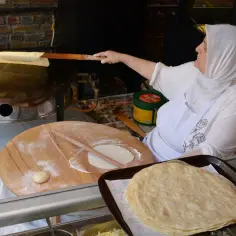 Kobieta w chuście przygotowuje tureckie jedzenie, smażąc cienkie placki na okrągłej płycie. Obok leżą gotowe placki i ciasto.