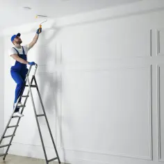 Mężczyzna w niebieskim kombinezonie stoi na drabinie, malując sufit wałkiem.