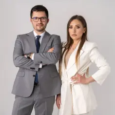 Para prawników w garniturach, mężczyzna w szarym, kobieta w białym, na jasnym tle.