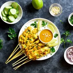 Pyszne satay z kurczaka na patyczkach, podane z sosem orzechowym, limonką, ogórkiem i posiekaną natką.