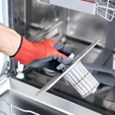 Ręka w rękawicy wyjmuje filtr zmywarki. Regularne czyszczenie filtra to klucz do świeżości naczyń, dlatego odświeżacze do zmywarek są zbędne jeśli dbasz o czystość filtra.