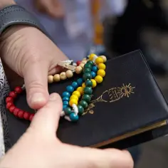 Kolorowy różaniec i czarna książeczka z symbolem IHS. W rękach trzymane są 10 przykazań Bożych.