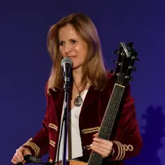 Wokalistka w bordowym żakiecie z ozdobnymi sznurami, z gitarą i mikrofonem, jakby śpiewała w piwnicy pod baranami.