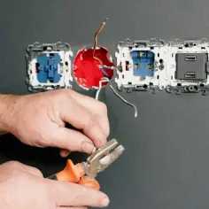 Ręce majsterkowicza przygotowują przewody do podłączenia gniazdka. Widać narzędzia i instalację elektryczną.