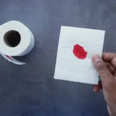 Dłoń trzyma biały papier toaletowy z niewielką plamką krwi. Obok leży rolka papieru.