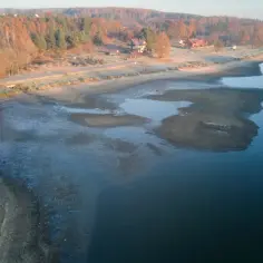 Widok na zalew brody z odsłoniętym dnem, otoczony jesiennym lasem i domkami.