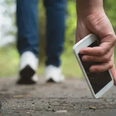 Dłoń podnosi biały telefon z ziemi na ścieżce. W tle widać nogi idącej osoby. To może być pomocne w lokalizacji telefonu.