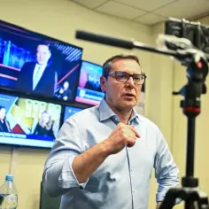 Prezes TVP w okularach, gestykuluje przed kamerą. W tle monitory z programami informacyjnymi.