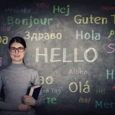 Kobieta z książką na tle tablicy z napisem "HELLO" i powitaniami w różnych językach. Czy to początek nauki top 10 najtrudniejszych języków świata?