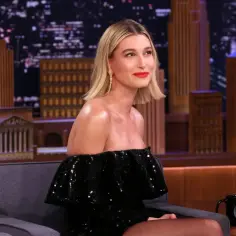 Hailey Bieber w ciąży, w czarnej, błyszczącej sukience, siedzi w studiu telewizyjnym.