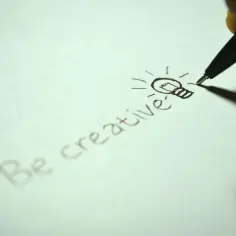Dłoń pisze długopisem "Be creative" i rysuje żarówkę, symbolizującą pomysł. To inspiracja w stylu popularnonaukowym.