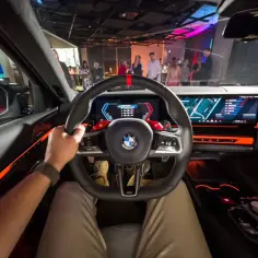 Wnętrze BMW M5 2025: dłoń na kierownicy, nowoczesne ekrany i pomarańczowe podświetlenie tworzą futurystyczny kokpit.