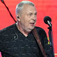 Adam Nowak śpiewa do mikrofonu, ubrany w czarną koszulę, z gitarą na ramieniu.