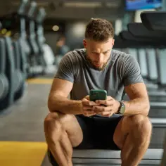 Mężczyzna w siłowni sprawdza telefon, szukając informacji o tym, co spala najwięcej kalorii. Tabela z ćwiczeniami jest w jego myślach.