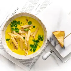 Aksamitna zupa neapolitańska z makaronem penne i natką pietruszki, podana na gazecie. Obok kawałek parmezanu i serowar.