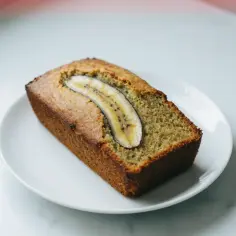Chlebek bananowy z airfryera, idealnie wypieczony, z plasterkiem banana na wierzchu, prezentuje się apetycznie na białym talerzyku.