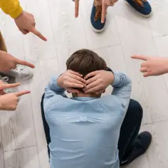 Chłopiec skulony na podłodze, otoczony przez palce wskazujące na niego. To obraz **bullyingu**, który sprawia, że czuje się samotny i przygnębiony.