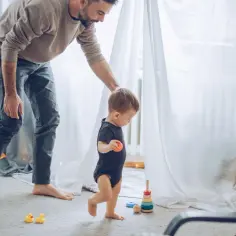 Tata pomaga synkowi, kiedy dzieci zaczynają chodzić. Na dywanie stoją kaczuszki i zabawki.