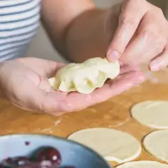 Dłonie formują pieroga z ciasta na pierogi bez jajka. Na blacie leżą okrągłe placki z ciasta i miseczka z wiśniami.