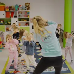 Wesołe zabawy dla dzieci w przedszkolu. Nauczycielka prowadzi zajęcia taneczne, a maluchy naśladują jej ruchy, tworząc radosną atmosferę.