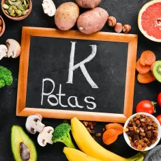 Tablica z napisem "K Potas" otoczona produktami bogatymi w potas: banany, ziemniaki, pomidory, awokado, brokuły, suszone morele, rodzynki, migdały, pestki dyni, orzechy, kiwi, grejpfrut. Pomaga zapobiegać hipokaliemii.