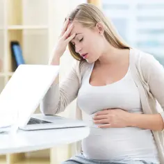Zmęczona ciężarna z dłońmi na czole i brzuchu, czuje kołatanie serca w ciąży przy laptopie.
