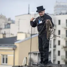 Kominiarz w cylindrze i czarnym stroju trzyma na dachu liny.