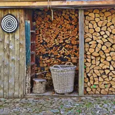 Drewno kominkowe ułożone w szopie, obok kosz i tarcza do rzutek. Widać różne sposoby, jak układać drewno kominkowe, by było suche i gotowe do palenia.