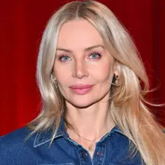 Agnieszka Woźniak-Starak w dżinsowej koszuli, z nowym partnerem u boku, prezentuje się promiennie na tle czerwonych zasłon.