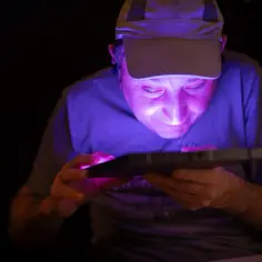 Mężczyzna w czapce z daszkiem pochyla się nad tabletem, którego ekran oświetla jego twarz fioletowym blaskiem. To jego osobista wystawa w ciemności.